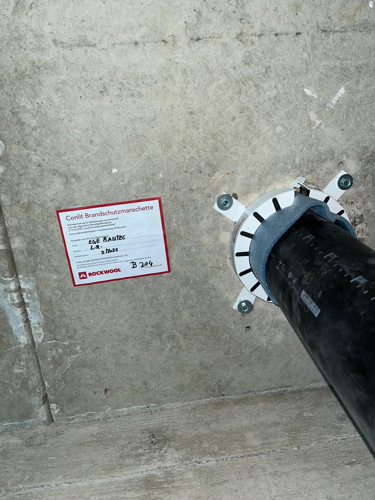 Brandschutzmanschette an einer Rohrdurchführung in einer Betonwand, daneben das ausgefüllte Kennzeichnungsschild mit Firma, Monteur, Jahr und Bauteilposition